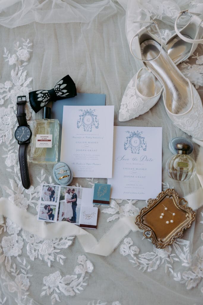Pepper Pavilion flat lay, with invitation suite, rings, bride's shoes, bow tie, blue ring box, groom's watch, perfume, cream ribbon.